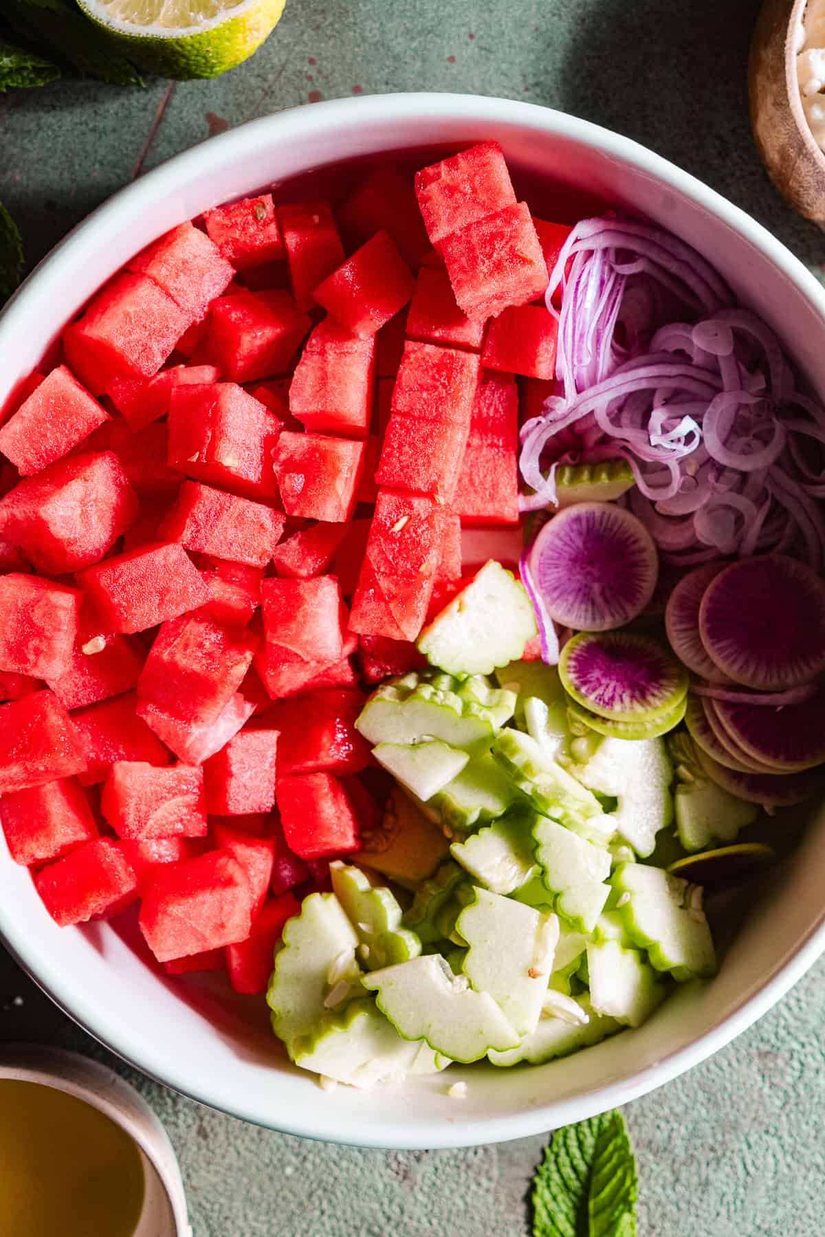Prepped fresh ingredients - watermelon, cucumber, red onion, and radish - for watermelon feta salad in a bowl.